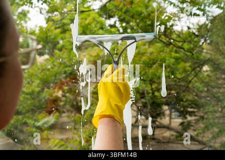 Femme dans des gants jaunes utilisant une raclette de nettoyage avec lavage d'un nettoyage de fenêtre dans sa maison. gros plan Banque D'Images