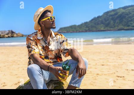 Jeune homme profitant d'une boisson rafraîchissante tout en portant des lunettes de soleil et un chapeau de paille, profitant du soleil sur une belle plage Banque D'Images