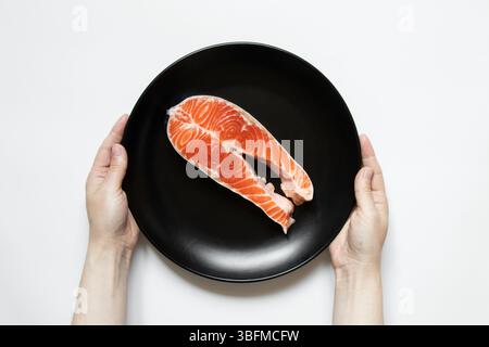 Un morceau de saumon cru repose dans une assiette noire et les mains de la jeune fille tiennent l'assiette sur un gros plan blanc Banque D'Images
