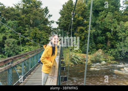 Jeune femme asiatique en veste jaune souriante et debout sur un pont suspendu en bois au-dessus de la rivière qui coule parmi la forêt tropicale au lac Matheson, Nouvelle-Zélande Banque D'Images