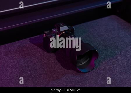 Un appareil photo numérique moderne noir sans miroir avec un grand objectif fixé, placé sur un fond sombre avec de subtils reflets de néon rose et bleu. Banque D'Images