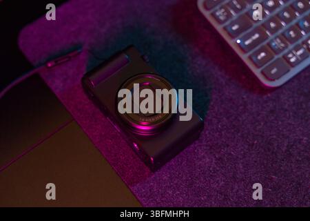 Plan macro détaillé d'un objectif compact d'appareil photo numérique, reflétant les néons roses et violets ambiants sur une surface texturée sombre. Cette image est parfaite Banque D'Images