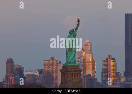 La Statue de la liberté lève son flambeau alors que la pleine lune se lève derrière elle, brillant au-dessus de la skyline de New York à l'heure dorée. Banque D'Images
