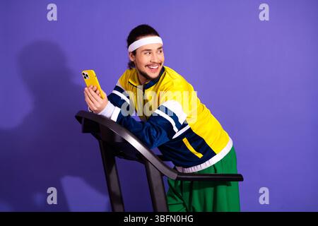 Jeune homme athlétique tenant smartphone tout en se tenant debout sur le tapis roulant sur fond violet, portant des vêtements de sport rétro Banque D'Images