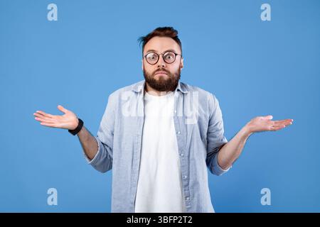 Qui sait. Homme barbu dans des lunettes haussant les épaules sur fond bleu studio Banque D'Images