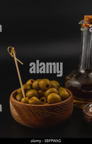 Un bol en bois rempli d'olives vertes se trouve à côté d'une bouteille en verre d'huile d'olive. Le riche bois brun contraste avec les olives vertes vibrantes dans un dimly Banque D'Images