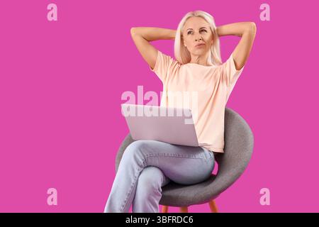 Femme mature réfléchie avec ordinateur portable assis dans un fauteuil sur fond violet Banque D'Images