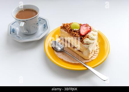 Une tranche de gâteau à la crème avec garnitures de fruits servi avec une tasse de thé au lait isolé sur blanc. Banque D'Images