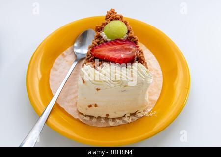 Gros plan d'une tranche de gâteau à la crème garnie de fraises et de raisin vert servie sur une assiette jaune isolée sur blanc. Banque D'Images