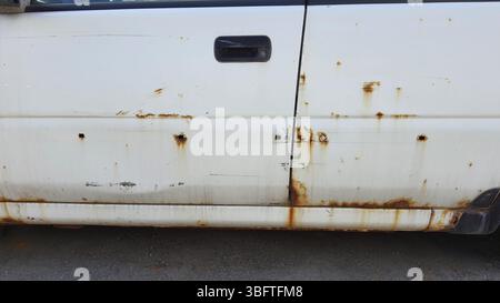 Vue rapprochée d'une porte de voiture blanche rouillée, montrant la corrosion et les dommages visibles, soulignant l'impact du temps et de la négligence sur le véhicule endommagé Banque D'Images