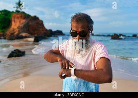 Homme ethnique senior avec la barbe grise sur la plage utilise une montre intelligente et des écouteurs pour le fitness. Les hommes âgés aiment la technologie pour la formation, le bien-être. Le vieil homme est entré Banque D'Images