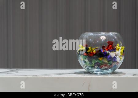 Un bol en verre transparent rempli de bonbons colorés emballés individuellement, assis sur une surface à motifs en marbre blanc. Un arrière-plan flou et rayé foncé Banque D'Images