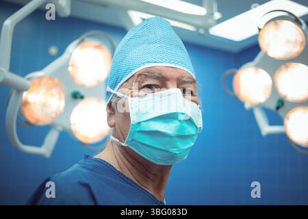 Chirurgien masculin senior debout dans la salle d'opération sous des lumières chirurgicales suspendues portant masque et casquette Banque D'Images