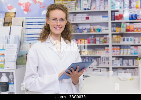 Pharmacienne de sexe féminin portant une blouse de laboratoire tenant un comprimé souriant au comptoir de la pharmacie avec des boîtes de médicaments Banque D'Images