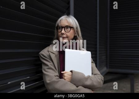 Une femme d'affaires d'âge moyen avec des lunettes se tient debout contre un mur sombre, tenant un ordinateur portable. Elle a l'air réfléchie et confiante, habillée dans un c élégant Banque D'Images