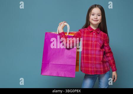 Isolé sur bleu, magnifique enfant brune caucasien en chemise rouge à carreaux et jeans bleus avec de longs cheveux tiennent des sacs en papier colorés Banque D'Images