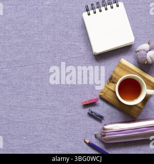 Bloc-notes, clip en bois, étui à crayons, tasse de thé, crayon de couleur, plante en coton sur fond de tissu violet. pose à plat, vue du dessus, espace pour copier. Travail et goujon Banque D'Images