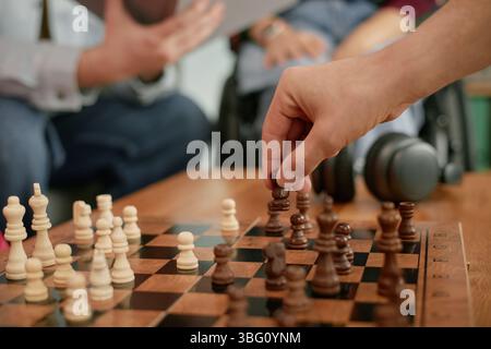 Homme caucasien handicapé jouant aux échecs avec un autre homme, gros plan des mains déplaçant des pièces d'échecs sur une planche de bois, fauteuil roulant visible en arrière-plan, célébration de l'anniversaire Banque D'Images