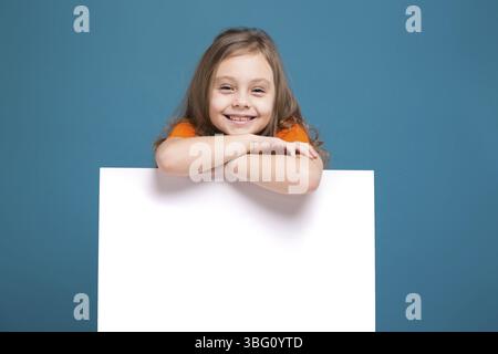 Isolé sur bleu, bel enfant caucasien avec de longs cheveux, en t-shirt orange et en Jean bleu montrent une affiche vide, se fie à l'affiche Banque D'Images