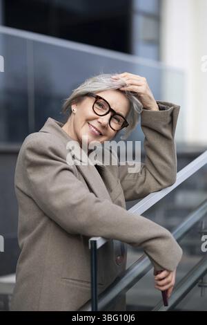 Une femme d'affaires d'âge moyen avec les cheveux gris se tient en toute confiance à l'extérieur, reposant sa main sur une balustrade en verre, souriant chaleureusement dans un env d'entreprise en plein air Banque D'Images