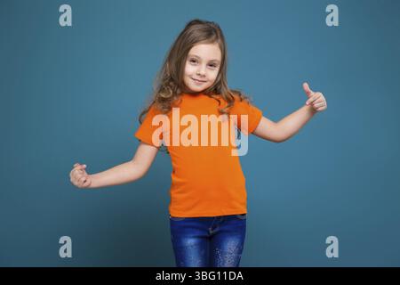 Isolé sur bleu, bel enfant caucasien avec de longs cheveux, en t-shirt orange et en Jean bleu, pointez vers elle-même, regardez la caméra Banque D'Images