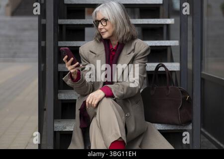 Une femme d'affaires caucasienne d'âge moyen s'assoit sur des escaliers extérieurs, élégamment habillée d'un manteau léger et d'un haut rouge vif, concentrée sur son smartphone pendant h Banque D'Images