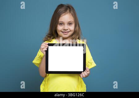 Isolé sur bleu, bel enfant caucasien avec de longs cheveux, en t-shirt jaune et pantalon noir montrent comprimé vide Banque D'Images