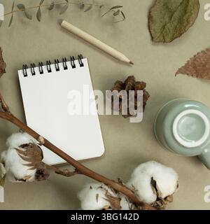 Concept hiver et automne. Carnet, tasse, crayon de couleur, plante en coton, feuilles sèches sur fond vert kaki. pose à plat, vue du dessus, espace pour les copies Banque D'Images