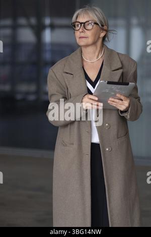 Une femme plus âgée vêtue d'un élégant manteau brun et de lunettes se tient à l'extérieur d'une structure contemporaine, tenant une tablette avec une expression concentrée sur elle Banque D'Images