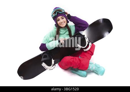 Isolé sur blanc, jolie fille caucasienne en tenue de ski violette, pantalon rouge, bottes de neige bleues et chapeau de laine violette assis jambes croisées avec neige noire Banque D'Images