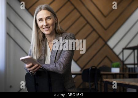 Une femme confiante avec de longs cheveux et un blazer s'assoit confortablement dans un espace de bureau contemporain, s'engageant avec son smartphone. Le mur géométrique en bois Banque D'Images