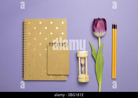 Bloc-notes, sablier, tulipe, crayon sur fond violet. plan à plat, vue de dessus, espace de copie. Lieu de travail et d'étude Banque D'Images