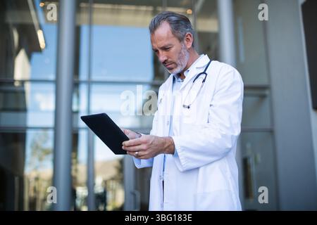 Médecin masculin principal debout à la porte de la clinique examinant la tablette et portant le stéthoscope, espace de copie Banque D'Images