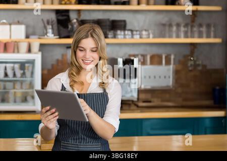 Femme portant un tablier vérifiant la tablette au comptoir du café et souriant près de la machine à espresso Banque D'Images