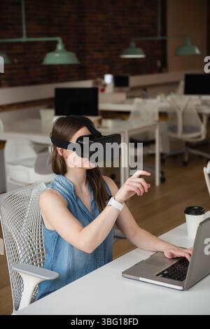 L'employée féminine porte un casque VR et est assise au bureau blanc, avec un ordinateur portable dans un bureau ouvert Banque D'Images