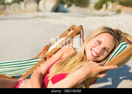 Femme s'inclinant sur une chaise de plage rayée sur la plage portant un bikini rouge tenant un chapeau de paille, espace de copie Banque D'Images