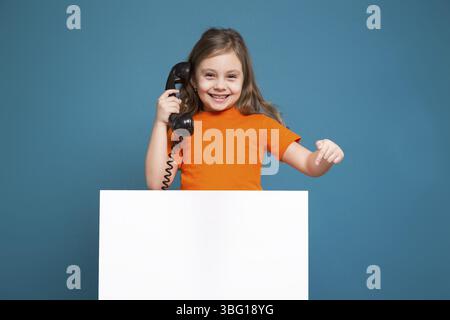 Isolé sur bleu, bel enfant caucasien avec de longs cheveux, en t-shirt orange et en Jean bleu montrent une affiche vide et tiennent le combiné du téléphone Banque D'Images