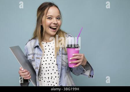 Image d'une joyeuse femme blonde européenne souriante et debout contre un mur bleu isolé avec un carnet argenté et un café à emporter dans les mains Banque D'Images