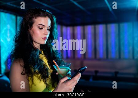 Femme portant une robe jaune tenant smartphone et verre martini près du banc dans le salon, espace copie Banque D'Images