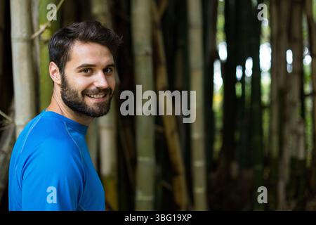 Homme debout et souriant en arrière sur l'épaule dans une bosquet de bambous portant un haut athlétique bleu, espace de copie Banque D'Images