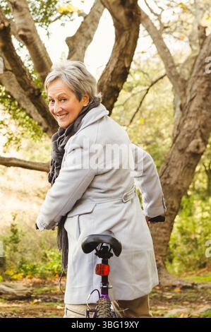 Femme senior debout dans un parc boisé tenant le guidon de vélo portant un manteau gris et une écharpe en tricot Banque D'Images