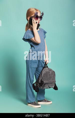 Isolé sur bleu, fille aux cheveux rouges dans des lunettes de soleil en forme d'étoile, salopette bleue et argentée, tenant un sac à dos brillant et parlant sur le noir Banque D'Images