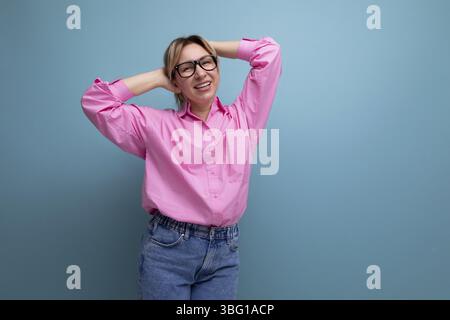 Dreamy positif jeune blond femme caucasienne modèle avec coiffure queue de cheval dans un look élégant composé de chemisier rose et de jeans Banque D'Images