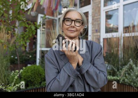 Une femme âgée bien soignée avec des cheveux gris et des lunettes est vêtue d'une veste grise et d'un Jean, tenant un smartphone dans sa main et attendant un appel Banque D'Images
