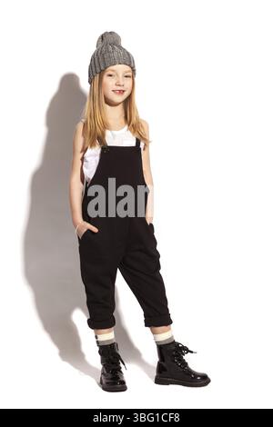 Isolé sur blanc, souriant fille aux cheveux rouges sans dent en salopette noire, t-shirt blanc, chapeau gris et chaussures laquées à lacets. les mains sont dans la poche Banque D'Images