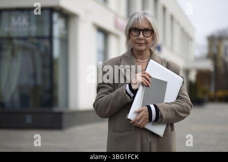 Une femme d'affaires âgée tient en toute confiance un portefeuille près de sa poitrine, vêtue d'un manteau élégant tout en se tenant debout à l'extérieur d'un bureau contemporain Banque D'Images