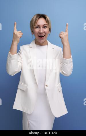 Joyeuse jolie blonde jeune femme européenne vêtue d'une veste blanche et robe pointe les doigts vers la publicité Banque D'Images