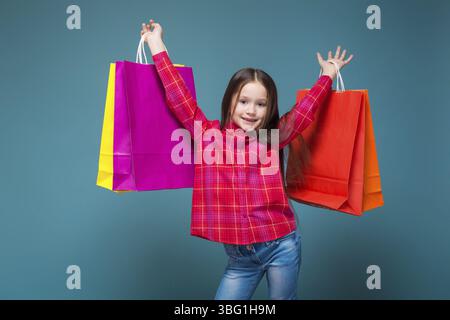 Isolé sur bleu, magnifique enfant brune caucasien en chemise à carreaux rouge et Jean bleu avec de longs cheveux tiennent des sacs en papier colorés, les mains vers le haut Banque D'Images