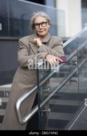 Une femme âgée élégante avec des cheveux gris et des lunettes se tient en toute confiance par une balustrade en verre dans un environnement urbain, profitant de l'ambiance diurne et s. Banque D'Images