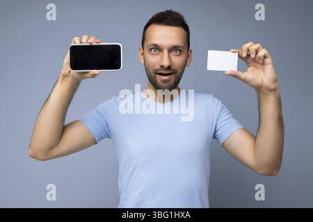 Photo d'un beau jeune homme peu rasé brunet portant une barbe t-shirt bleu de tous les jours isolé sur fond bleu tenue et utilisation téléphone mobile et Banque D'Images